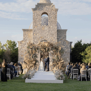 Bride and groom exchanging vows outside a stone chapel with floral arch.