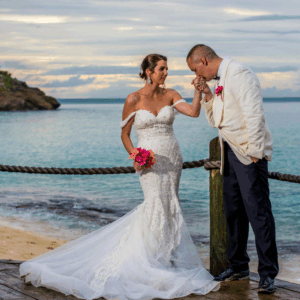 Groom kissing bride’s hand on a dock by the ocean at sunset.